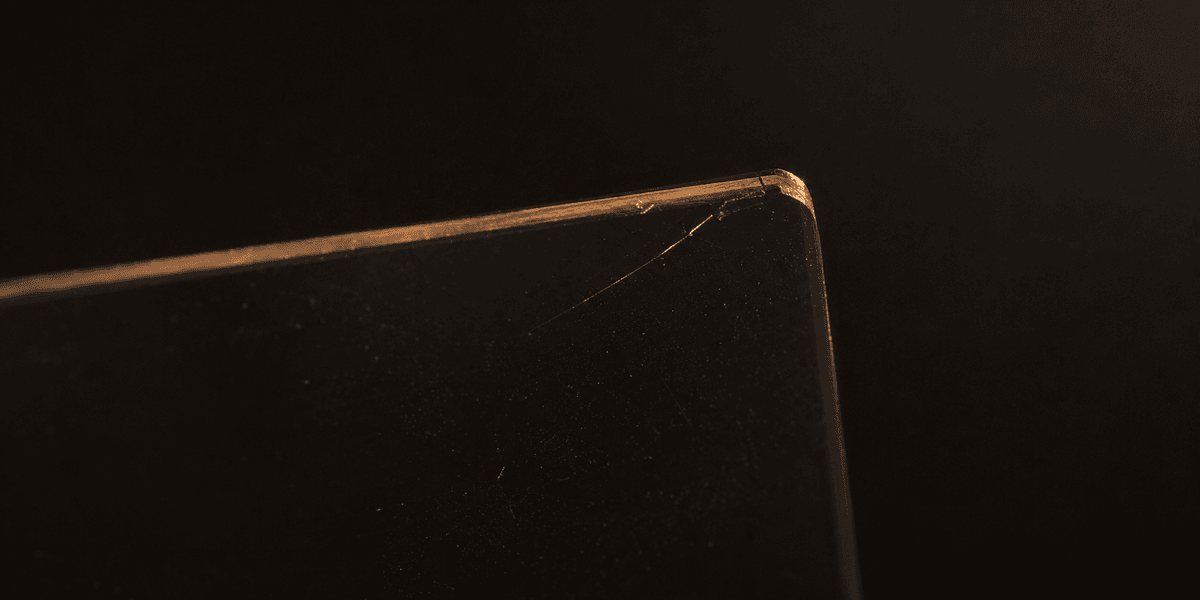 A close-up of a hairline crack along the edge of a dark glass panel, catching a thin line of light — representing the small, easy-to-miss flaw that quietly undermines an otherwise solid surface